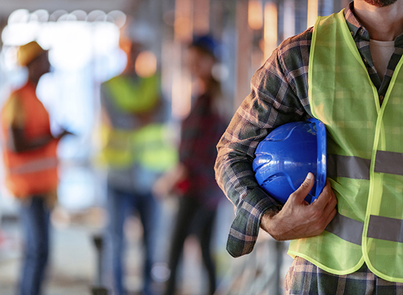 Man holding blue helmet close up. Construction man worker with office and people in background. Close up of a construction worker's hand holding working helmet.