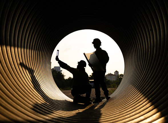 Construction workers and engineers are working efficiently inside a large metal pipe at a construction site. They are building a new water drainage system in the evening, just before sunset.
